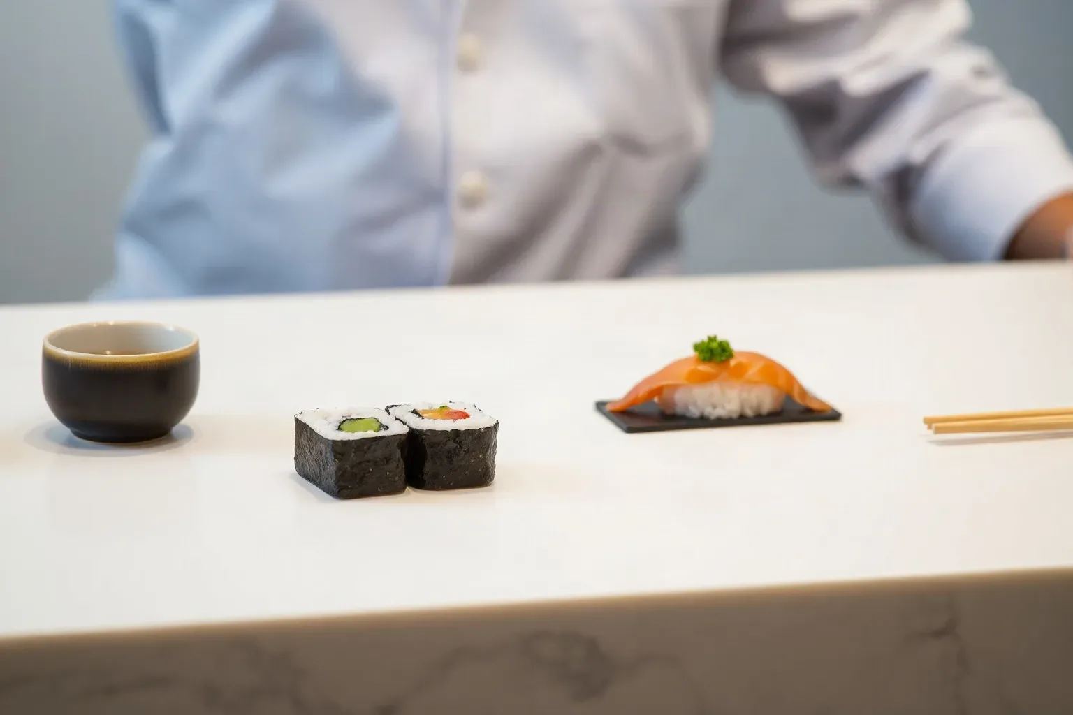 Two small ceramic cups beside a participant's own completed maki and nigiri pieces on a class preparation counter