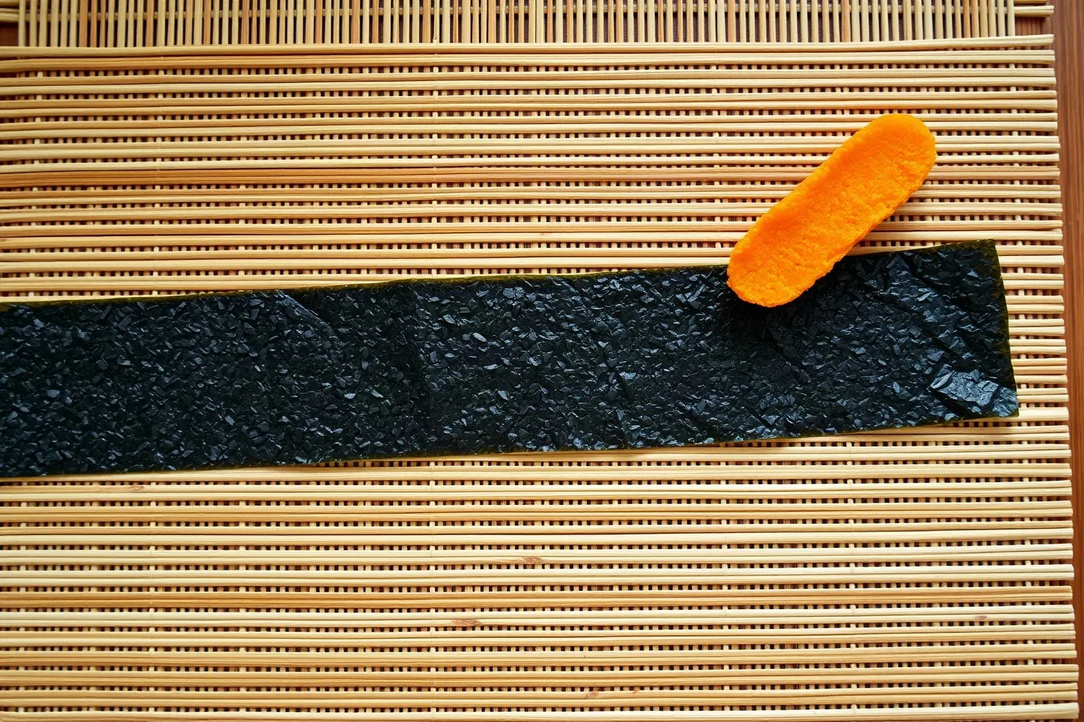 A bamboo rolling mat with a nori sheet laid flat and a strip of sweet potato tempura placed at the near edge ready for a